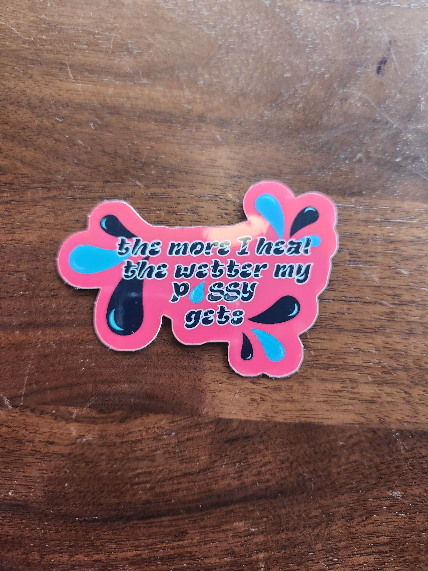 A pink sticker is the shape of a water splash, laying on a wooden table
The words are black and outlined in white. They say: "the more I heal the wetter my p-u-s-s-y gets"
In the corners are black and blue water droplets