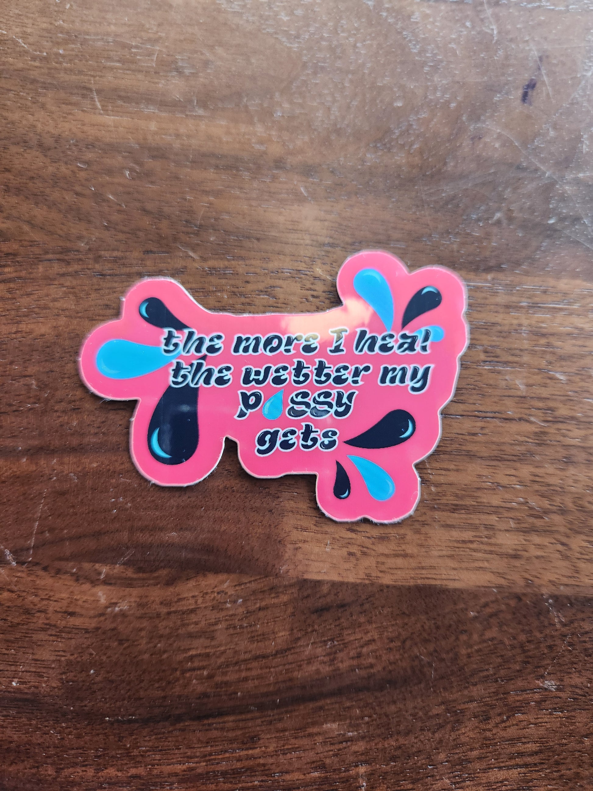 A pink sticker is the shape of a water splash, laying on a wooden table

The words are black and outlined in white. They say: "the more I heal the wetter my p-u-s-s-y gets"

In the corners are black and blue water droplets 